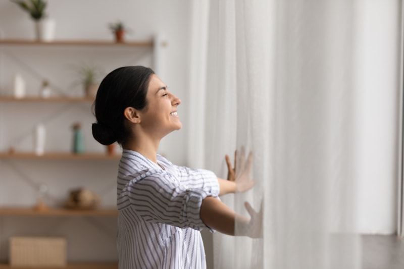 smiling woman opening up curtains in her home on a sunny day.