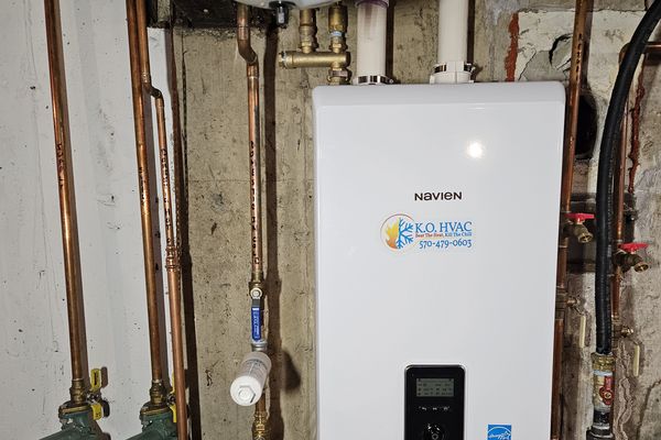 A Navien tankless water heater with a KO HVAC sticker on a basement wall, with copper piping
