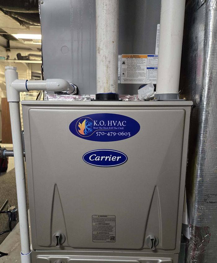 A Carrier gas furnace with the KO HVAC logo plate and white PVC venting, installed in a basement